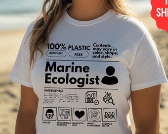 May include: White t-shirt with a black graphic design that says "100% Plastic Free Marine Ecologist". The graphic includes a person icon, a heart, a wave, and a book.