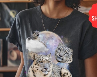 Könnte beinhalten: Ein schwarzes T-Shirt mit einem Grafikdruck von drei Schneeleoparden in einer Galaxie-Kulisse mit einem Vollmond. Die Schneeleoparden sind weiß mit schwarzen Flecken. Der Mond ist hellblau mit einem Galaxiemuster.