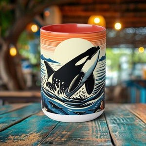 May include: A white ceramic mug with a red rim features a graphic of an orca whale jumping out of the water. The background is a sunset with orange, yellow, and blue colors.