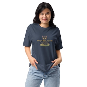 Könnte beinhalten: Ein dunkelblaues T-Shirt mit kurzen Ärmeln und einer Grafik eines kleinen schwarzen Katers, der auf einem grünen Grasfleck mit hohen Grashalmen im Hintergrund sitzt. Der Text "I Don't Bite... Usually" ist in goldener Schrift darüber gedruckt.