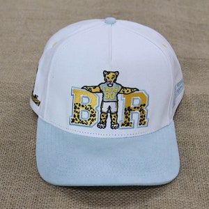 May include: A white baseball cap with a light blue brim. The cap features an embroidered design of a jaguar mascot with the letters "B R" in gold with a leopard print pattern. The cap has a light blue button on top.