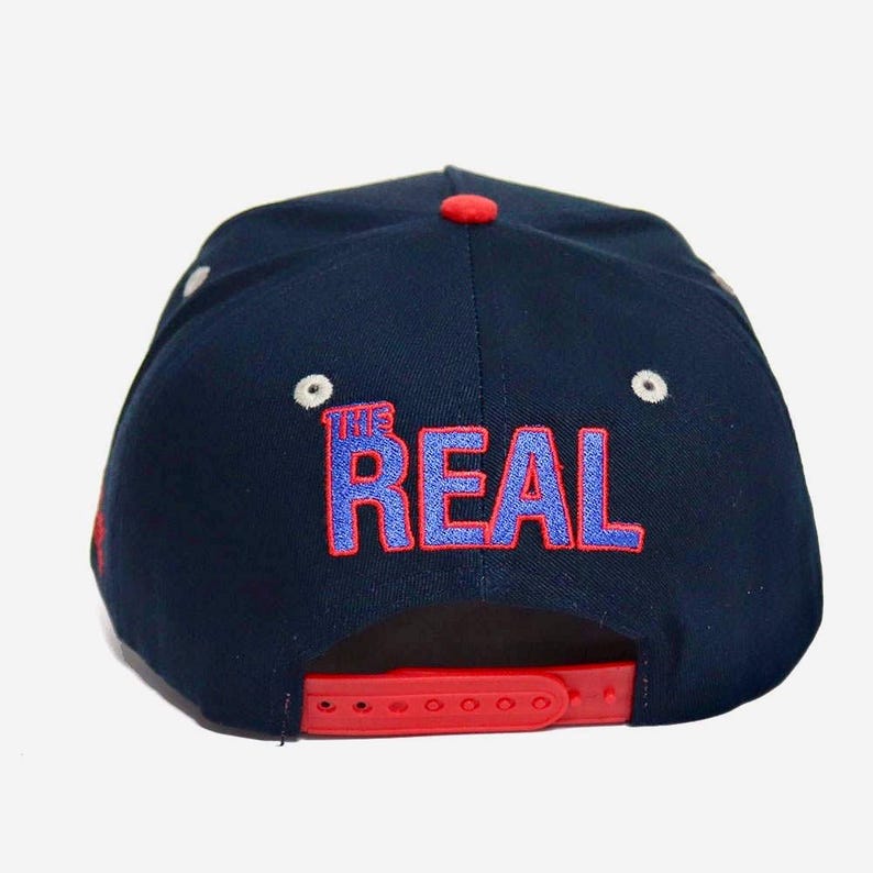 May include: Navy blue baseball cap with a red bill and adjustable strap. The back of the cap features the text "THE REAL" in blue and red embroidery. The cap has a red button on top and white eyelets.