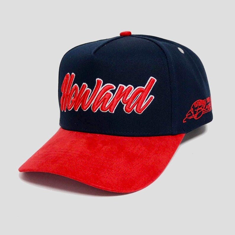 May include: A navy blue baseball cap with a red suede brim. The word "Howard" is embroidered in red script across the front. A small red logo is on the side of the cap. The cap has a red button on top.