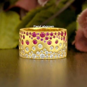 Peut inclure: Une bague en or avec une surface texturée, ornée de pierres précieuses rouges et blanches. La bague présente une bande de pierres rouges en haut, une section centrale avec des pierres dorées et blanches et une section inférieure avec des pierres blanches.