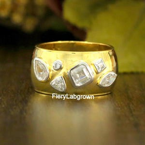 May include: A gold-colored ring with multiple clear, geometric-shaped stones. The ring has a wide band and the text "FieryLabgrown" is visible on the ring. The ring is set against a blurred background of flowers.