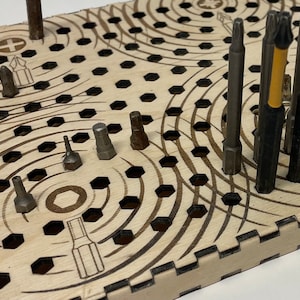Op de afbeelding: Een houten gereedschapsorganizer met een cirkelvormig patroon en meerdere gaten om verschillende schroevendraaierbits op te bergen. De organizer heeft een lichtbruine houtkleur en een zwart-wit ontwerp.