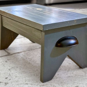 Op de afbeelding: Een grijze houten opstapje met een zwart metalen handvat. De opstapje heeft een verweerde afwerking en is gemaakt van massief hout.
