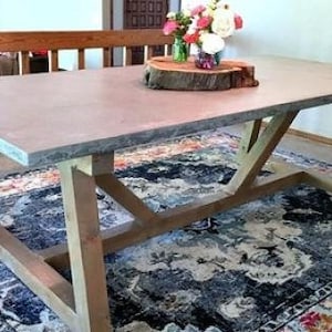 Puede incluir: Una mesa grande, rectangular de hormigón con un acabado gris claro y una base de madera. La mesa tiene un diseño simple y moderno y está soportada por cuatro patas que están conectadas por una abrazadera en forma de cruz. La mesa está sentada sobre una alfombra colorida con un patrón azul y blanco.