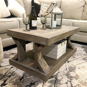 May include: A rustic wooden coffee table with a lower shelf. The table has a distressed brown finish and features a white wooden box on the lower shelf. Two wine glasses and a bottle of wine are on the table's surface.