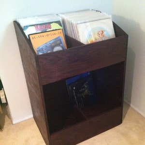 Könnte beinhalten: Ein dunkelbraunes Holzregal für Schallplatten mit zwei Fächern. Der obere Bereich enthält mehrere Vinylplatten in Hüllen, darunter eine mit dem Text "Misfits" und "at.the.drive.in". Der untere Bereich enthält ein paar Platten.