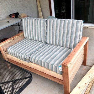 Op de afbeelding: Een houten buitenbank met een gestreept kussen. De bank is gemaakt van onbehandeld hout en heeft een eenvoudig ontwerp.