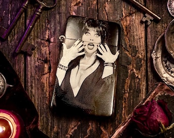 May include: Black and white lighter featuring a portrait of a woman with dark hair and dramatic makeup. The lighter is set against a dark, textured wooden surface, surrounded by antique keys and decorative elements. The image has a vintage, gothic aesthetic.