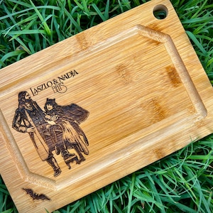 May include: A wooden cutting board with a dark brown engraved design of two figures and the text "Laszlo & Nadja Bats".