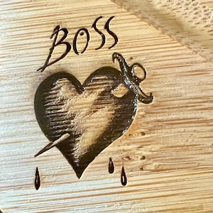 May include: A wooden cutting board with the word "BOSS" etched at the top. Below the word is a heart pierced by a dagger, with three teardrops. The design is burned into the light-colored wood.