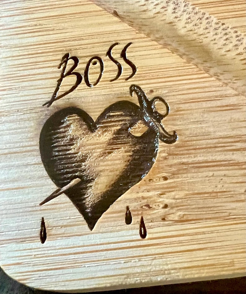 May include: A wooden cutting board with the word "BOSS" etched at the top. Below the word is a heart pierced by a dagger, with three teardrops. The design is burned into the light-colored wood.