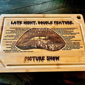 May include: A rectangular bamboo cutting board with a carved design. The design includes a large, detailed image of lips, surrounded by text. The words "LATE NIGHT, DOUBLE FEATURE" and "PICTURE SHOW" are also carved into the board.