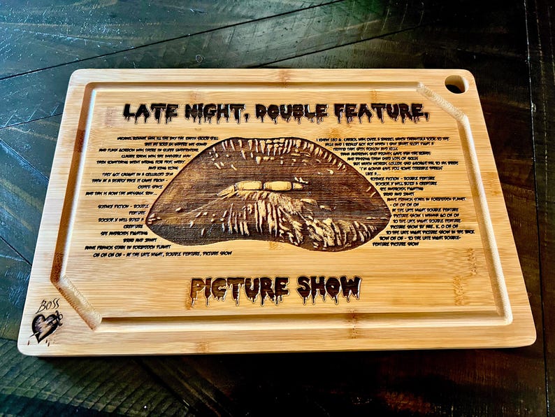 May include: A rectangular bamboo cutting board with a carved design. The design includes a large, detailed image of lips, surrounded by text. The words "LATE NIGHT, DOUBLE FEATURE" and "PICTURE SHOW" are also carved into the board.