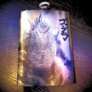 May include: A silver-toned metal flask featuring a detailed laser-etched graphic of the monster Godzilla on the front surface. The design includes Japanese kanji characters vertically aligned on the right side and a screw-top cap at the centre.