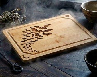 Puede incluir: Tabla de cortar rectangular de bambú con un diseño tallado de murciélagos en vuelo. La tabla tiene un canal para jugos y un agujero para colgar. Los murciélagos son de color marrón oscuro, con una luna creciente tallada.