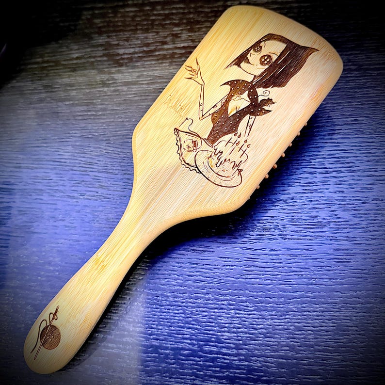 May include: A wooden hairbrush with a handle and a wide, rectangular head. The brush features a detailed, engraved illustration of a character with large eyes and a dark dress. The handle has a decorative design. The brush is made of light-colored wood.
