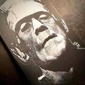 May include: Black and white illustration of Frankenstein's monster. The image is a close-up of the monster's face, with a prominent brow, stitched neck, and a bandaged head. The artwork has a vintage, classic horror movie aesthetic.
