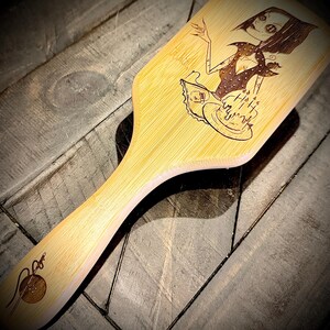 May include: A light-colored wooden paddle brush with a handle. The brush features a laser-etched design of a cartoon woman with a cake and the words "Happy Birthday". A needle and thread design is etched on the handle.