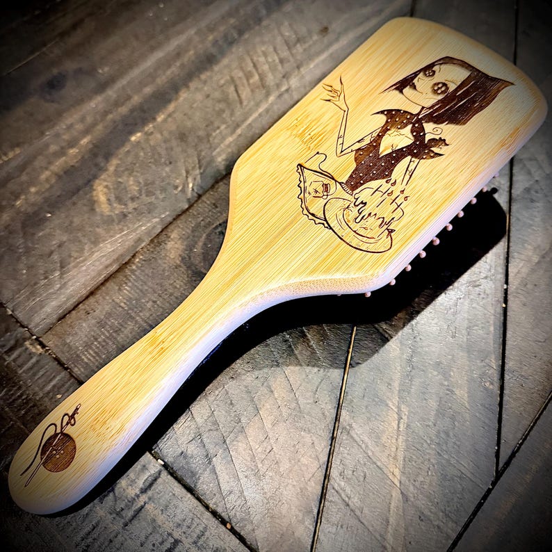 May include: A bamboo hairbrush with a laser-etched illustration of a cartoon character. The character is wearing a polka-dot dress and holding a cake. The brush has a handle and pink bristles.