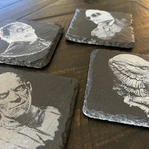 May include: Four black slate coasters with white etched images of classic horror movie monsters. The coasters feature the Creature from the Black Lagoon, the Mummy, Frankenstein's Monster, and the Wolfman.