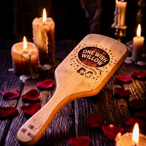 May include: A light-coloured wooden hairbrush with a flat, rectangular head and a long handle. The head features an engraved design with the text "ONE WISH WILLOW" and "AMAZE YOUR FRIENDS" above two faces. The handle is engraved with "ONLY ONE WISH".