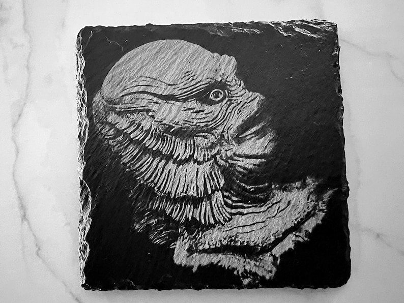 May include: Black and white image of a creature from the black lagoon on a slate coaster.