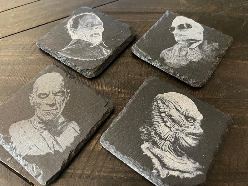 May include: Four black slate coasters with white images of classic horror movie monsters. The coasters feature the Creature from the Black Lagoon, the Invisible Man, the Wolfman, and the Mummy.