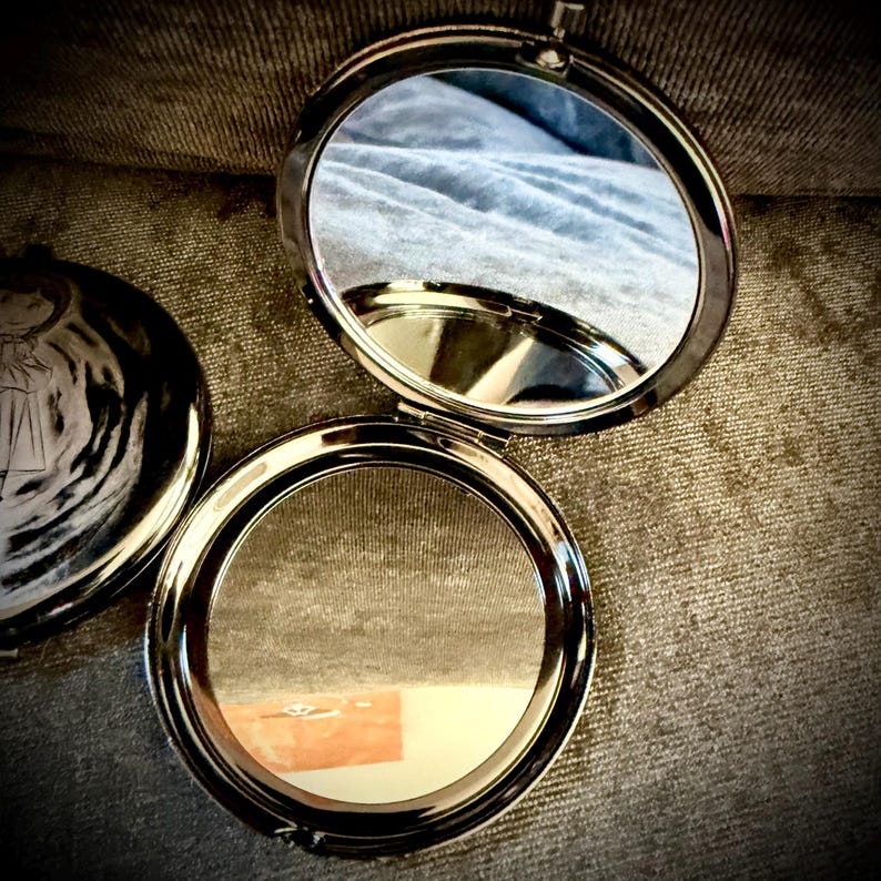 May include: Open, silver-toned compact mirror with a round shape. The mirror is reflecting a light blue surface. The compact is on a textured, brown surface. Another silver-toned compact is partially visible to the left.