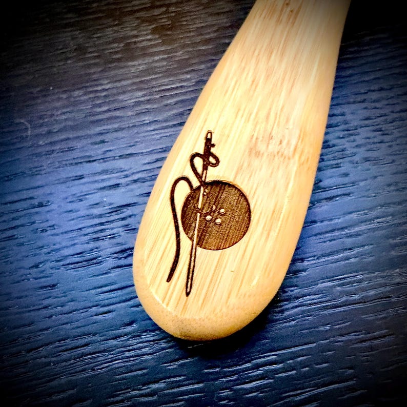 May include: A wooden sewing tool with a laser-etched design. The design features a button, needle, and thread. The tool is made of light-colored wood and has a smooth, curved shape. The background is a dark blue.