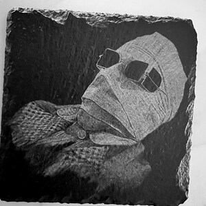May include: Black and white image of a person's face with bandages and sunglasses on a black slate coaster.