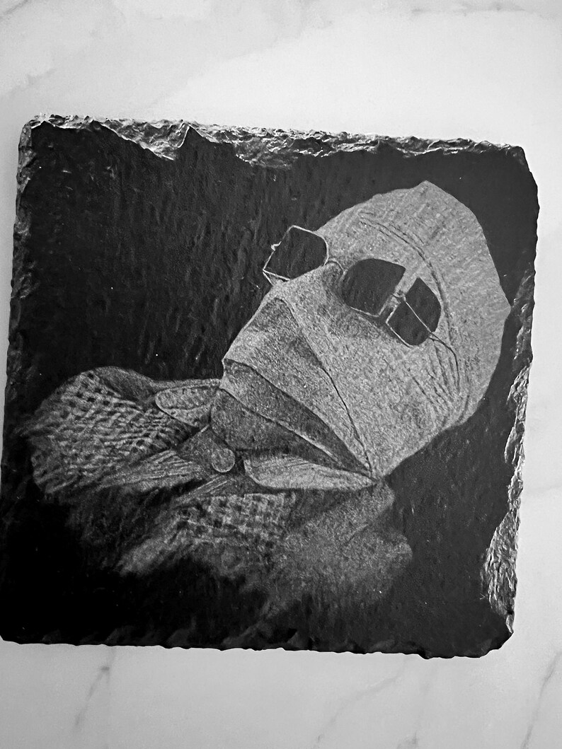 May include: Black and white image of a person's face with bandages and sunglasses on a black slate coaster.