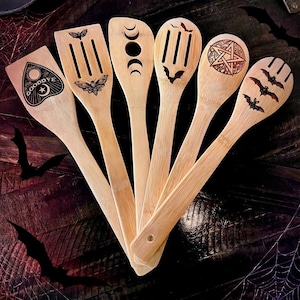 May include: Set of six bamboo kitchen utensils with engraved designs. Includes a spatula with a planchette design, a slotted spatula with a moth, a spoon with crescent moons, a slotted spoon with a bat, a spoon with a pentagram, and a slotted spoon with bats.