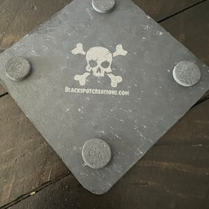 May include: A gray slate coaster with a white skull and crossbones design. The coaster has four gray felt pads on the bottom. The text "BLACKSPOTCREATIONS.COM" is printed on the coaster.