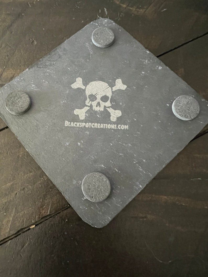 May include: A gray slate coaster with a white skull and crossbones design. The coaster has four gray felt pads on the bottom. The text "BLACKSPOTCREATIONS.COM" is printed on the coaster.