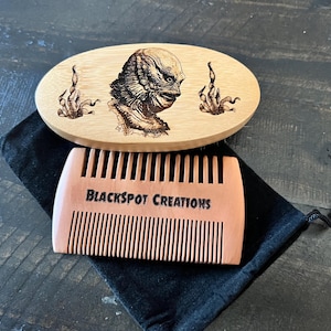 May include: A wooden beard comb and a wooden beard brush with a Creature from the Black Lagoon design. The comb is peach-coloured with the text "BLACKSPOT CREATIONS". The brush is oval-shaped.