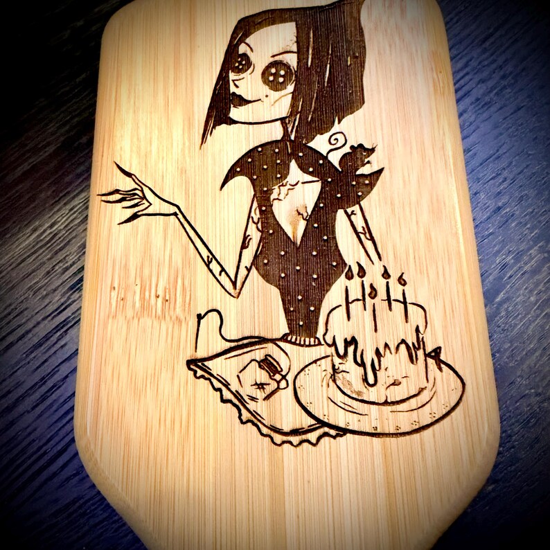 May include: A wooden cutting board with a pyrography design. The design features a stylized illustration of a character holding a cake with candles. The character has button eyes and a dark dress with polka dots. The board has a light wood grain.