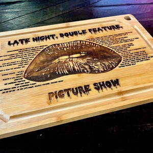 May include: A rectangular bamboo cutting board with a carved lip design. The words "LATE NIGHT, DOUBLE FEATURE" and "PICTURE SHOW" are carved into the wood. The board has a juice groove and a heart with the word "Boss" on the corner.