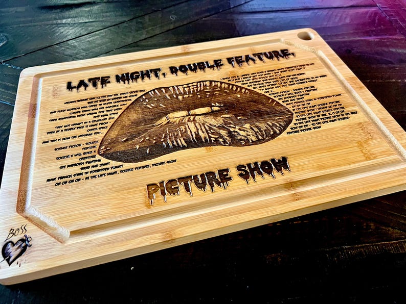 May include: A rectangular bamboo cutting board with a carved lip design. The words "LATE NIGHT, DOUBLE FEATURE" and "PICTURE SHOW" are carved into the wood. The board has a juice groove and a heart with the word "Boss" on the corner.