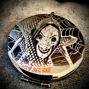 May include: A round, silver compact mirror featuring a black and white graphic of a character with a cracked face and a sinister smile. White spiderwebs are on the edges of the mirror, adding to the spooky theme.