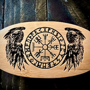 Könnte beinhalten: Ein ovaler Untersetzer aus Holz mit einem schwarzen und weißen gravierten Design. Das Design zeigt einen Kompass mit Runen um den Außenrand und zwei Flügeln auf jeder Seite.