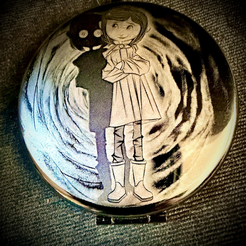 May include: A round, silver-toned pendant featuring an engraved illustration of a girl in a coat and boots, with a dark silhouette of a figure behind her. The background has a swirling, textured design.