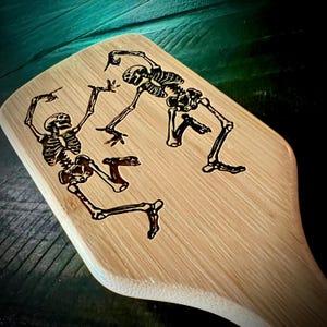May include: A light-colored wooden paddle with a dark, etched design of two dancing skeletons. The skeletons are in dynamic poses, with arms and legs outstretched. The paddle has a curved shape and a handle.