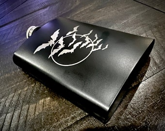 May include: A black metal flask with a silver design of bats flying around a circle.
