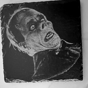 May include: Black and white drawing of a man's face on a black slate coaster. The man has a pale complexion and is looking up with a surprised expression. His mouth is open and his teeth are visible. The drawing is done in a realistic style.