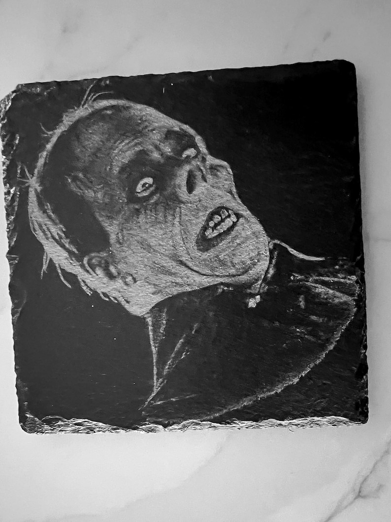 May include: Black and white drawing of a man's face on a black slate coaster. The man has a pale complexion and is looking up with a surprised expression. His mouth is open and his teeth are visible. The drawing is done in a realistic style.
