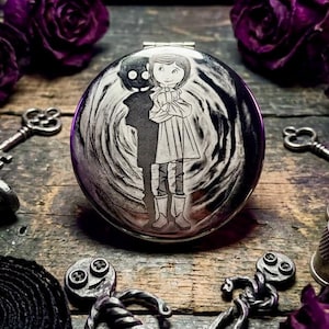 May include: A round, silver compact mirror with a detailed engraving of a girl and a shadowy figure. The design is set against a swirling background. The mirror is surrounded by keys, thimbles, and purple roses on a wooden surface.